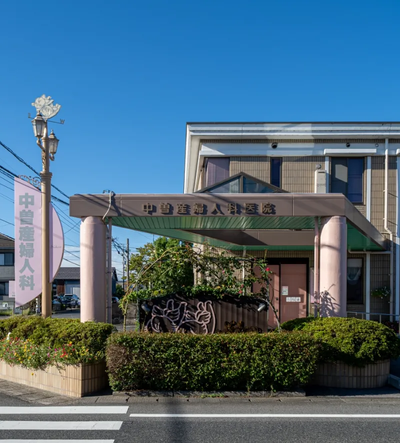 青空の下、低層の建物の正面入口を写した写真。入口上部には『中澤産婦人科医院』と書かれた看板があり、その手前には植え込みがある