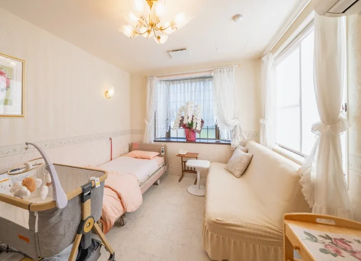 明るい日差しが差し込む、ピンクとクリーム色を基調とした広々とした病室。ベッドが2台、ソファ、そして左端にはベビーベッドが見える。