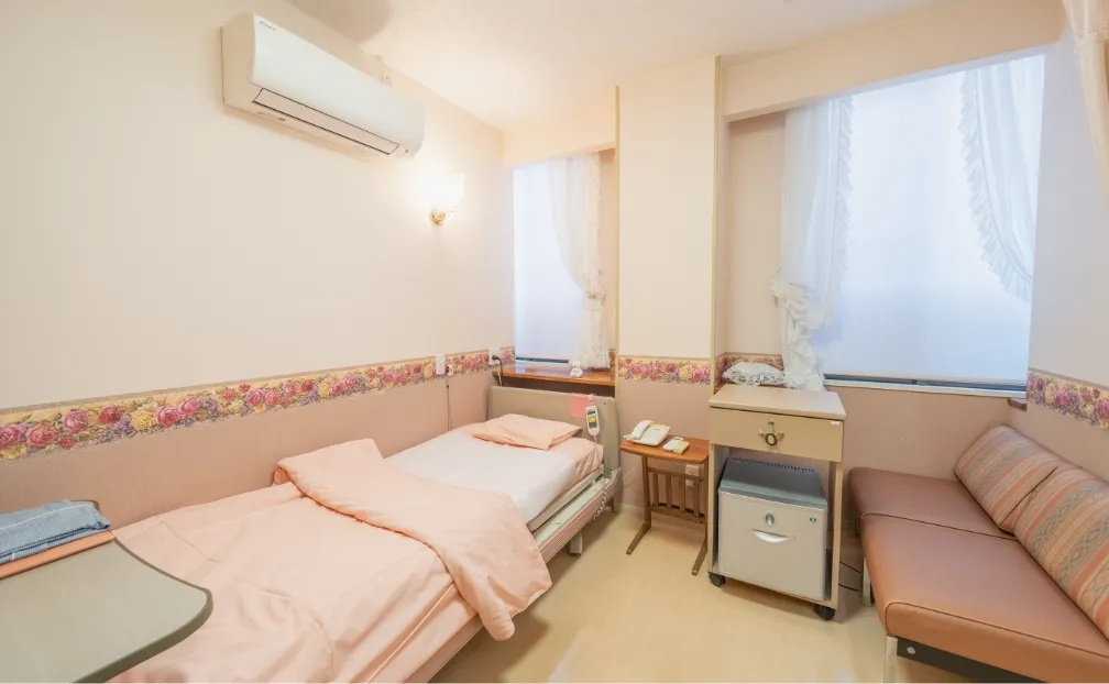 通常室の内部。ピンクの壁と花柄のボーダーが特徴的な、明るいシングルベッドの病室。壁にはエアコンがあり、窓際には小型のソファとサイドテーブルが置かれている。
