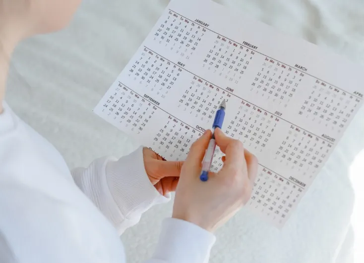女性がカレンダーまたは基礎体温表をペンで指し示しているクローズアップ。月経周期の管理や排卵日の特定、または診療日の確認の様子。