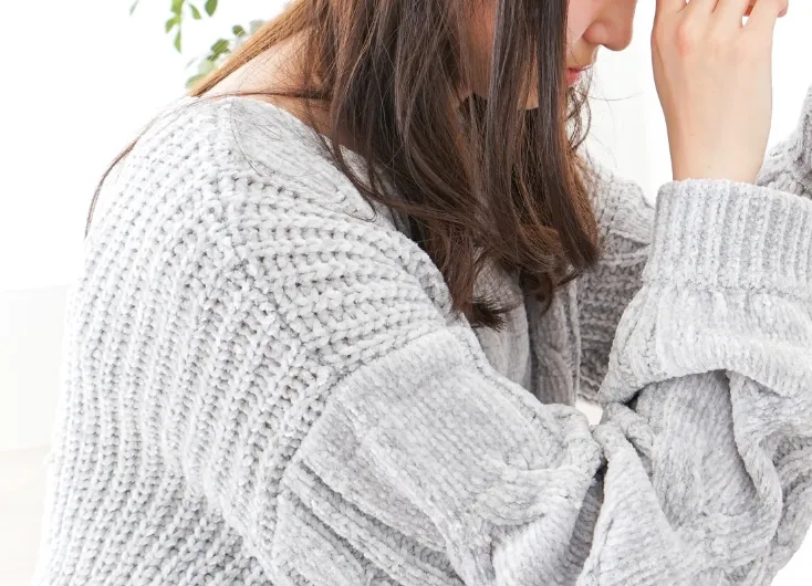 グレーのセーターを着た女性が顔を両手で覆い、肩を落としているクローズアップ。精神的なストレス、PMS（月経前症候群）、または落ち込みなどの心理的な不調を表現している。