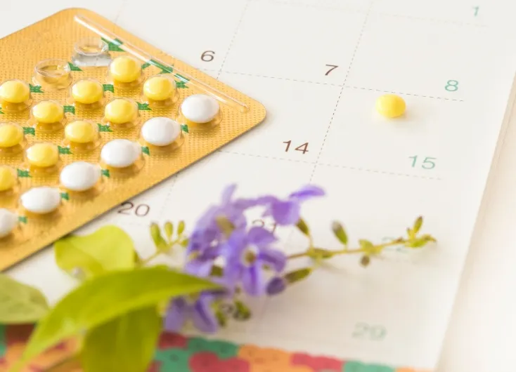 カレンダーの上に置かれた経口避妊薬（ピル）のシートと、紫色の小花。避妊相談やピル処方をイメージさせるクローズアップ。
