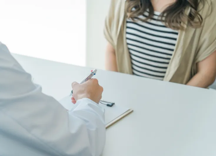 白い制服を着た医師が、向かいに座る患者を前にしてカルテに書き込みをしている様子。望まない妊娠や緊急避妊に関する相談、または診察のイメージ。